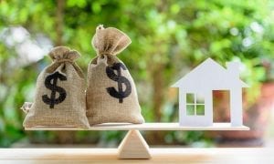 Two money bags and a paper model of house balancing on wooden scale