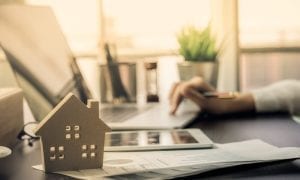 Wooden model of home in front of womans hand browsing laptop with documents on desk