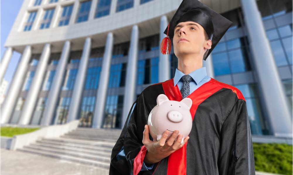 Young male student gazing into distance holding piggy-bank in front of university building
