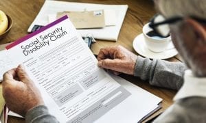Male senior with grey hair and glasses holding social security disability claim form in hand on table