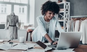 Young businesswoman reaching over paper filled desk to access laptop