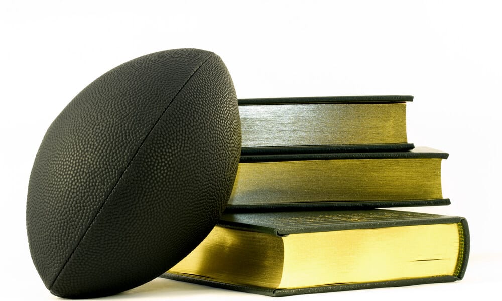 Black football leans against three, gold edged books, unifying symbols of sports, academic, and learning achievement